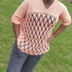 May include: A woman wearing a peach crochet top with a lacy pattern and dark blue pants. She is standing in a grassy field.