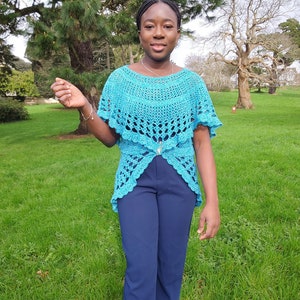 May include: A woman wearing a teal crochet top with a high-low hemline. The top has a delicate lace pattern and is worn over a pair of dark blue pants.