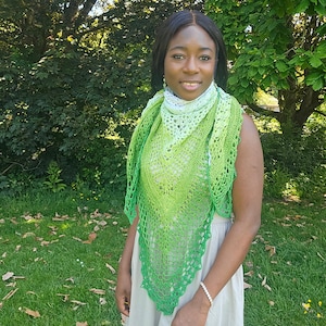 May include: A triangular crochet shawl featuring an ombre color gradient that transitions from white at the neckline to light green and deep forest green at the pointed hem. The shawl has an intricate openwork lace pattern with a scalloped edge.
