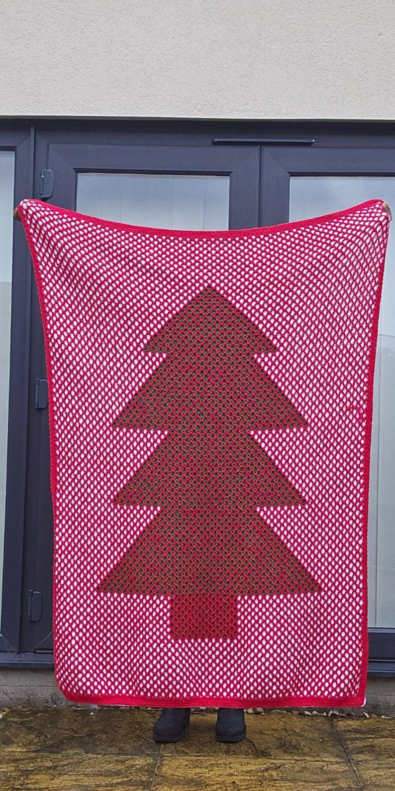 May include: A red and white crocheted blanket featuring a Christmas tree design in the center. The tree is a darker shade of red. The blanket is held up in front of a door.