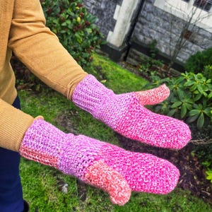 May include: A pair of handmade mittens in shades of pink, purple, and coral. The mittens are knitted with a textured stitch and feature a thumb. The background includes green grass and foliage.