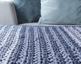 Può includere: Una coperta in maglia grossa blu e grigia con un motivo testurizzato. La coperta è piegata e posizionata su un divano bianco con due cuscini blu.