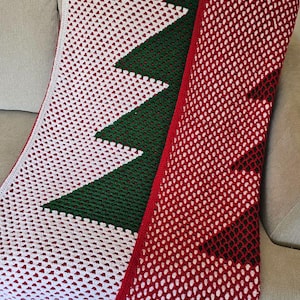 May include: A handmade crochet blanket featuring a Christmas tree design. The blanket has a white background with a green tree and a red border. The blanket is draped over a gray couch.