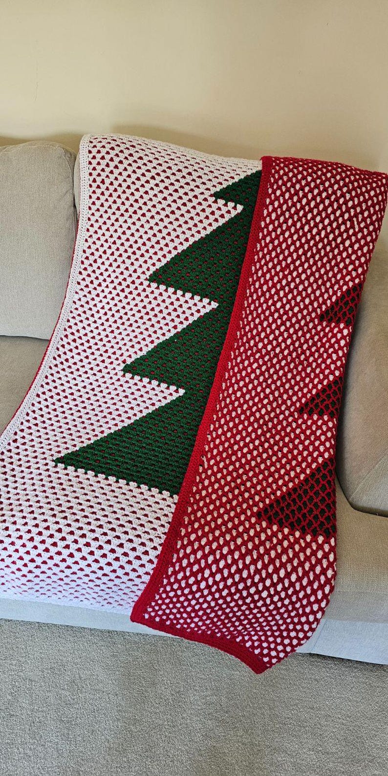 May include: A handmade crochet blanket featuring a Christmas tree design. The blanket has a white background with a green tree and a red border. The blanket is draped over a gray couch.