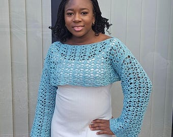 May include: A light blue crochet bolero with long sleeves. The bolero has an open, lacy design. The garment is worn over a white dress. The sleeves are flared at the wrist. The bolero is a handmade item.