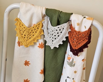 May include: Three crocheted dishcloths hanging on a white rack. The dishcloths are in shades of yellow, beige, and orange. The dishcloths are decorated with a scalloped edge.