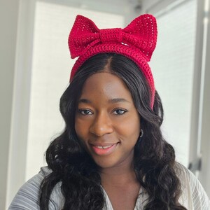 May include: A red crocheted headband with a large bow on top. The headband is a vibrant shade of red and appears to be handmade. The bow is prominently displayed, adding a decorative element to the accessory.