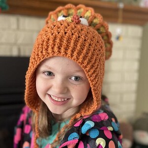 May include: A young person wearing a crocheted orange and brown turkey hat with a white pom-pom on top. The hat has a brown crocheted brim and a white crocheted beak. The person is wearing a black and pink polka dot jacket.