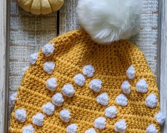 May include: A yellow crocheted beanie with white polka dots and a white faux fur pom pom. The beanie is sitting on a white wooden surface with two small yellow gourds.