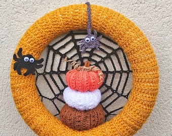 Könnte beinhalten: Ein gehäkelter Halloween-Kranz mit einem schwarzen Spinnennetz, drei gestapelten Kürbissen und zwei schwarzen Spinnen mit Glitzern.