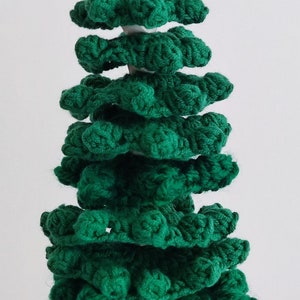May include: A green crocheted Christmas tree ornament with a wooden top. The tree is made of multiple layers of crocheted fabric, creating a layered and textured look.
