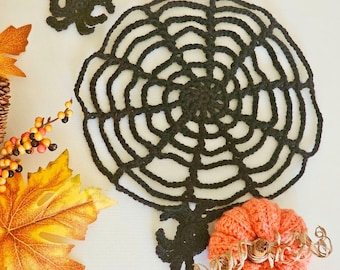 May include: A black crocheted spiderweb with a black crocheted spider on a white background. There is a small orange crocheted pumpkin in the lower right corner of the image.