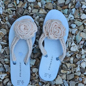 May include: A pair of white flip-flops with crocheted pink flower embellishments. The flip-flops have the text "PRIMARK" and size information: "UK/IRL 5/6 EUR 38/39 USA 7/8".