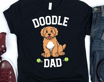 May include: Black t-shirt with a graphic of a tan Doodle dog and the words "DOODLE DAD" in white. Two green tennis balls are on either side of the word "DAD". The shirt is made of a soft material.