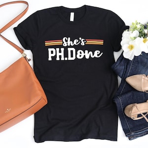 May include: Black t-shirt with the text "She's PH.Done" in white, with horizontal stripes in orange and yellow. A brown purse, blue jeans, and beige flats are also visible.