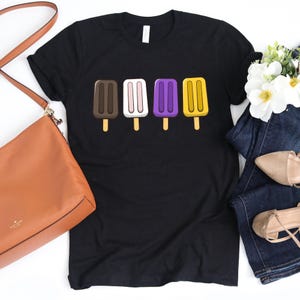 May include: Black t-shirt featuring a row of four colorful popsicle illustrations. The popsicles are brown, pink, purple, and yellow, each with a wooden stick. The shirt is paired with a brown purse, jeans, and shoes.