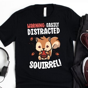 May include: Black t-shirt with a cartoon squirrel graphic. The squirrel is brown and holding an acorn. Text on the shirt reads "WARNING: EASILY DISTRACTED SQUIRREL!" in red and white.