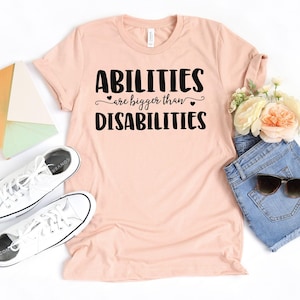 May include: A light pink t-shirt with the text "ABILITIES are bigger than DISABILITIES" in black. The shirt is styled with white sneakers, denim shorts, sunglasses, and flowers. The shirt is made of a soft material.