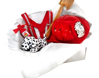 Pode incluir: Uma caixa branca cheia de um capacete de bombeiro vermelho, um machado de brinquedo, um extintor de brinquedo vermelho e branco, um brinquedo de pelúcia dálmata preto e branco e suspensórios vermelhos.