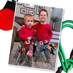 May include: Two young boys wearing red shirts and blue and yellow patterned belts with various toys attached. The boys are standing in a living room with a brown couch and a green rug.
