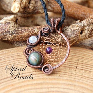 Può includere: Ciondolo acchiappasogni in filo di rame con cordino nero. Il ciondolo presenta un design a ragnatela con una gemma viola al centro e altre due gemme. Il testo "Spiral Roads" è visibile.