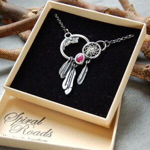 Puede incluir: Un collar de plata con un dije de atrapasueños, un círculo con una piedra preciosa rosa y un diseño de plumas. El collar está en una caja beige con el texto "Spiral Roads" impreso en la parte inferior.