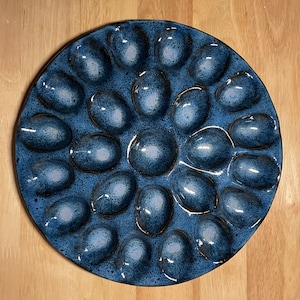 May include: A round, blue ceramic egg serving platter with 18 egg-shaped indentations. The platter has a glossy finish with a speckled pattern. The background is a light wood surface.