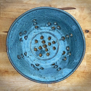 May include: A blue ceramic colander with a circular shape and a textured surface. The colander has a central circle of holes and a ring of smaller holes around the edge.