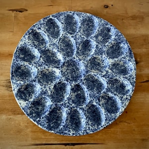 May include: A round, blue and white speckled ceramic egg serving platter. The platter has 24 egg-shaped indentations. The speckled pattern is a mix of dark blue and white. The platter is on a wooden surface.