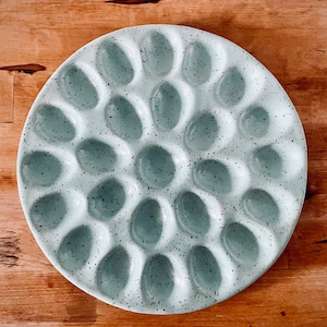 May include: A round, speckled, ceramic serving platter with 19 oval indentations. The platter is a pale blue-green color with dark gray speckles. The design is ideal for serving deviled eggs or other appetizers. The platter sits on a wooden surface.