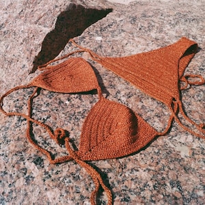 May include: A crocheted bikini top in a burnt orange color. The top is made of a fine yarn and has a triangular shape. The top is lying on a rocky surface.