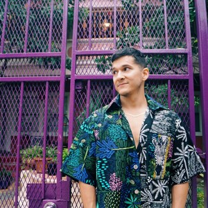 May include: A person wearing a black and green patterned shirt with a white collar and short sleeves. The shirt has a variety of colorful floral and geometric designs. The person is standing in front of a purple metal gate.