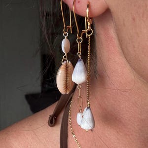 Può includere: Orecchini dorati con design a conchiglia e perline. Un orecchino presenta una conchiglia beige e una perla bianca. L'altro ha un guscio bianco e una catena dorata con dettagli di conchiglie bianche. Gli orecchini sono appesi all'orecchio di una persona.
