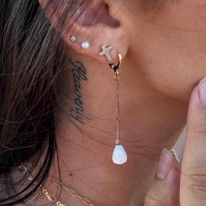 May include: Close-up of an ear with multiple earrings. One earring is a gold hoop with a cross charm, and a long chain ending in a white teardrop-shaped stone. Other earrings include a small stud and a pearl. A gold necklace is also visible.
