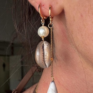 May include: Gold hoop earrings with a pearl, a seashell, and a white cone-shaped charm. The earrings hang from a gold chain. The seashell is a light brown color.