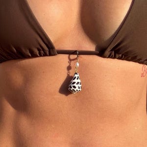 May include: A brown bikini top with a seashell pendant. The pendant is a teardrop shape, with a black and white pattern, and a small pearl. The pendant hangs from a gold-colored hoop.