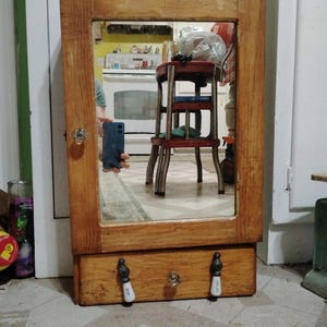 May include: A rectangular wooden wall cabinet featuring a central mirror with a clear glass knob. Below the mirror is a single drawer equipped with two white and black ceramic handles and a central clear glass knob, all finished in a natural wood tone.
