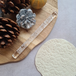 May include: A clear, textured rolling pin with a detailed floral pattern, next to a round of clay with a matching design. The image includes decorative pinecones and a small gourd on a wooden board.