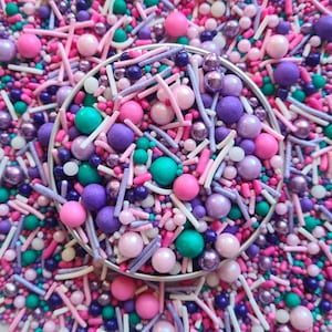 May include: A close-up of a selection of colourful sprinkles, including pink, purple, teal, and white. The sprinkles are in a small metal container and are ideal for decorating cakes, cupcakes, and other desserts.