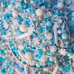 May include: A close-up of a silver bowl filled with blue and white sprinkles, including snowflake shapes, stars, and small balls.