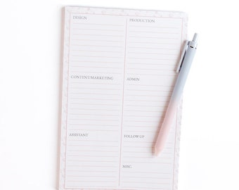 May include: A white notepad with a floral border and lined pages. The notepad is divided into sections labeled "Design", "Production", "Content/Marketing", "Admin", "Assistant", "Follow Up", and "Misc.". A pink and gray pen is lying on top of the notepad.