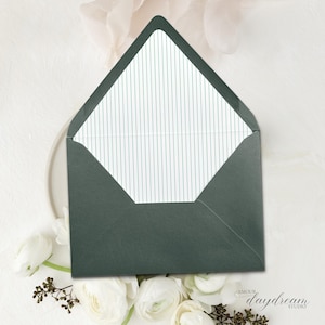 May include: A dark green envelope with a white interior featuring thin, vertical teal stripes. The envelope is open, revealing the striped interior. White flowers and greenery surround the envelope. The text "AMOUR daydream STUDIO" is visible.