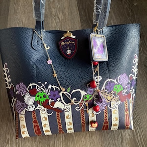 Puede incluir: Bolso tote azul marino con un borde decorativo con flores moradas, calaveras verdes y manzanas rojas. El bolso tiene una cadena dorada con dijes y un parche que dice "Dormfore".