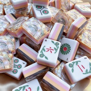 May include: A collection of rectangular mahjong tiles featuring a white top surface with green floral engravings and red Chinese characters. The sides of the tiles display layered bands of pink, orange, and brown, while some tiles show a pearlescent finish.