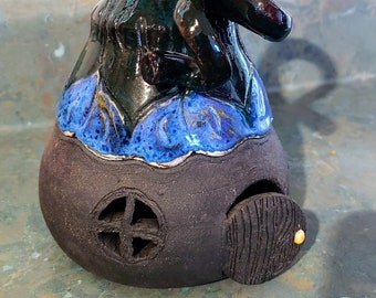 May include: A ceramic gnome house with a black and green glazed roof and a blue and green glazed base. The house has a small door and a window.