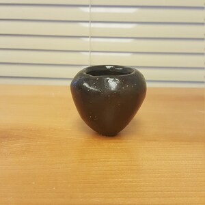 Puede incluir: Una pequeña maceta de cerámica negra de forma redonda. La maceta está sobre una superficie de madera.