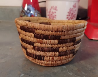 Puede incluir: Una pequeña cesta tejida con un patrón marrón y beige. La cesta está hecha de materiales naturales y tiene una forma redonda.
