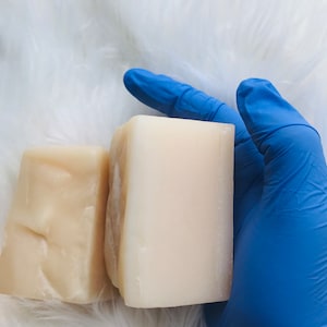 以下が含まれることがあります： 青いラテックス手袋に、白とベージュの2つの石鹸が握られています。石鹸は長方形で、表面は滑らかです。