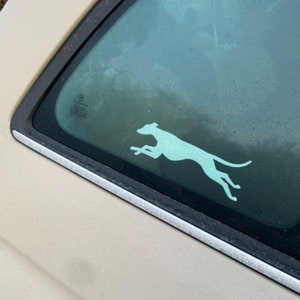 Puede incluir: Una pegatina de silueta blanca de un galgo corriendo en la ventana trasera de un coche beige.