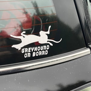 Peut inclure: Une silhouette blanche d'un l&eacute;vrier courant, avec le texte "Greyhound On Board" en dessous, sur une vitre de voiture noire.